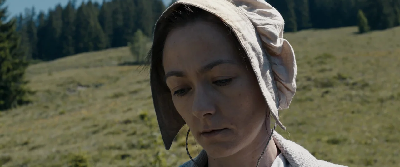 Hagazussa (2017) movie still: closeup — A woman wears a traditional linen bonnet in an open, grassy alpine meadow. She keeps her head bowed while…