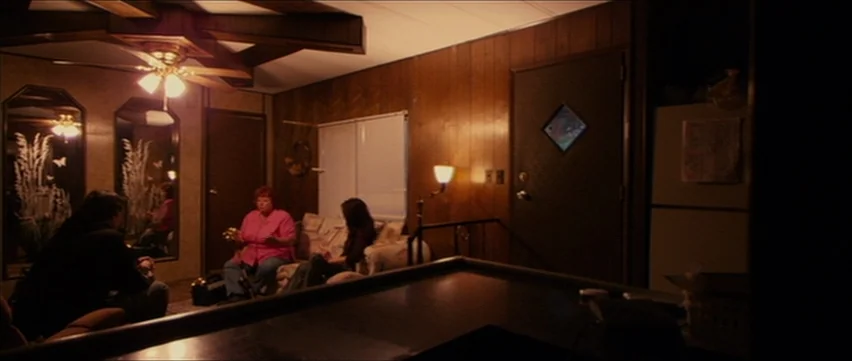 Bubble (2005) movie still: wide — A dimly lit mobile home living room features wood-paneled walls and a ceiling fan. Three people are seated in the…