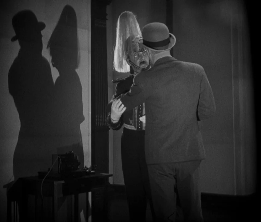 The Threepenny Opera (1931) movie still: medium — A sparsely furnished room features a wooden table with a small electronic device. A man in a suit…