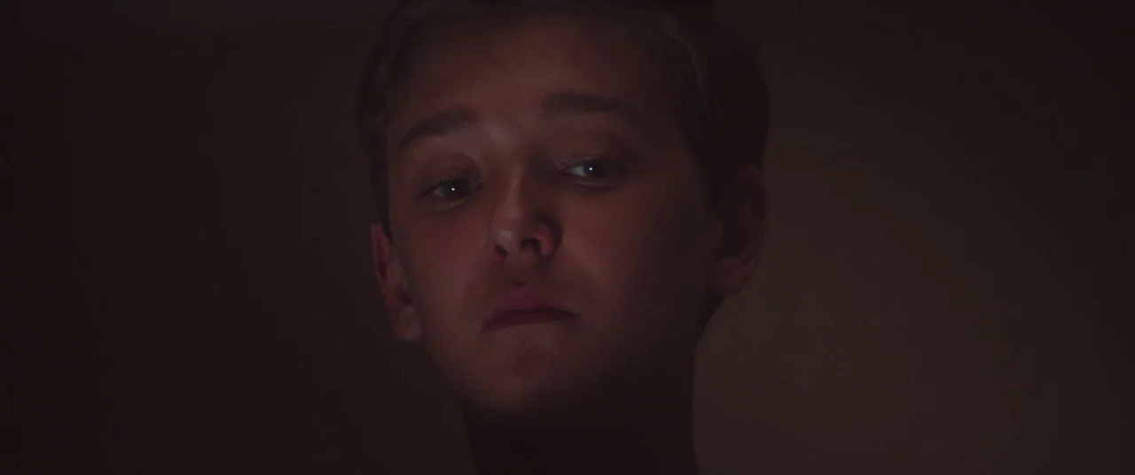 The Strange Ones (2017) movie still: closeup — The frame shows a dimly lit interior space with a dark, indistinct background. A young male subject…