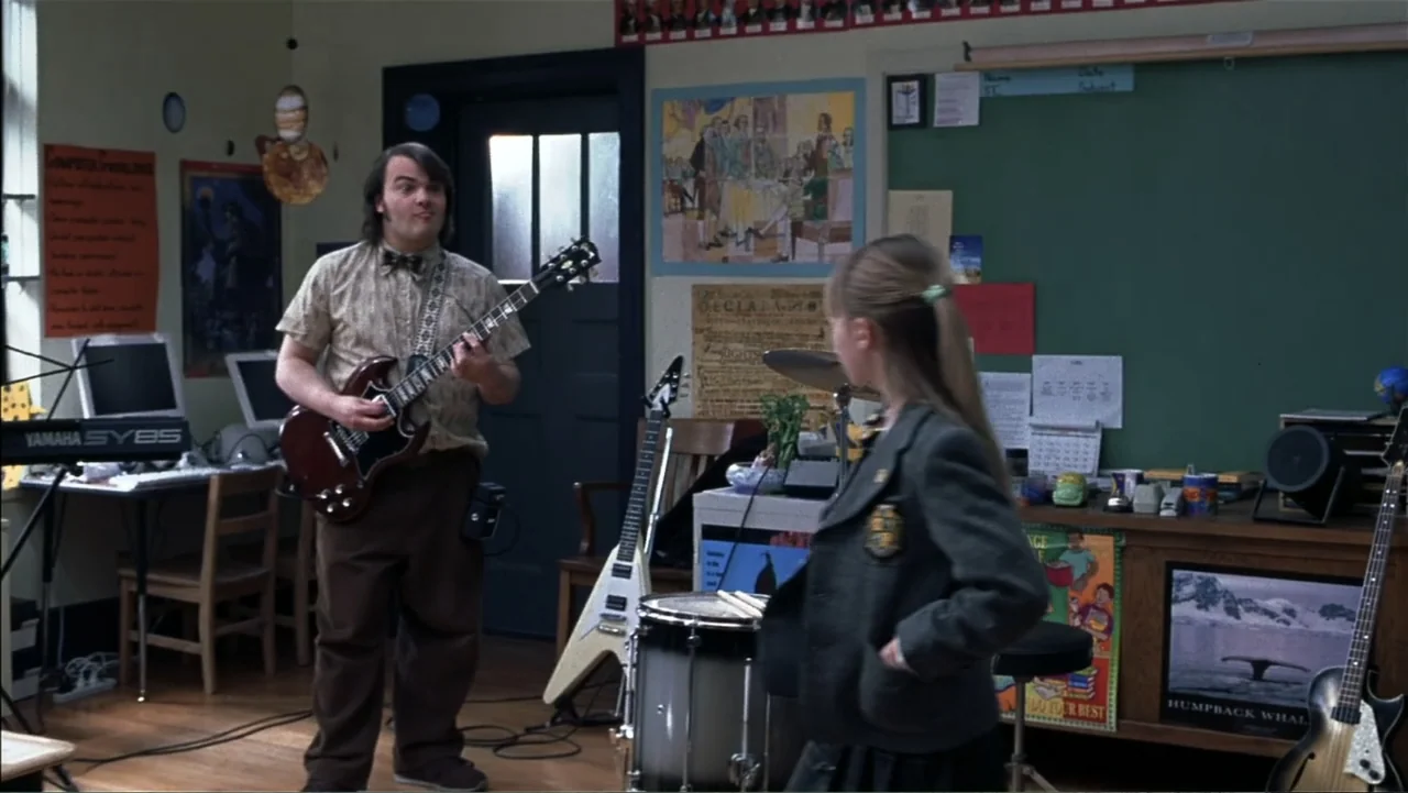 School of Rock (2003) movie still: fullbody — A cluttered school classroom features posters, computers, and multiple musical instruments scattered…