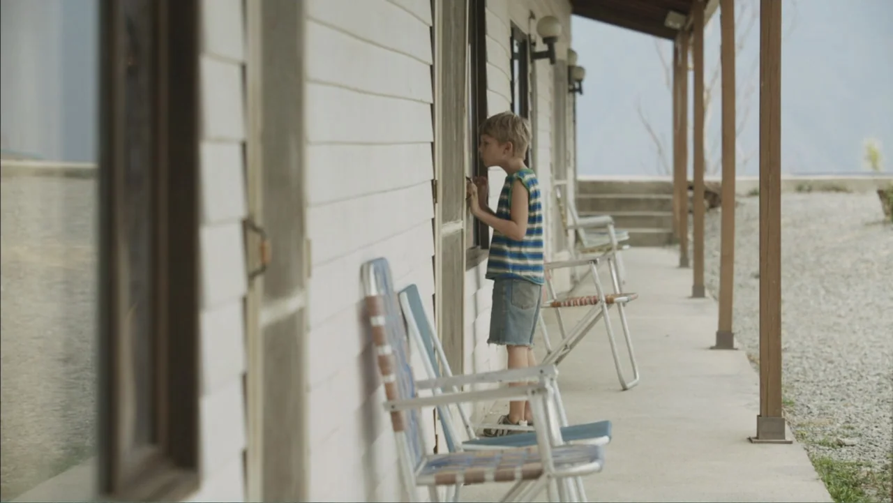 Boy (2010) movie still: fullbody — A long exterior porch with white siding lines a building with several glass doors. A young boy stands near a…