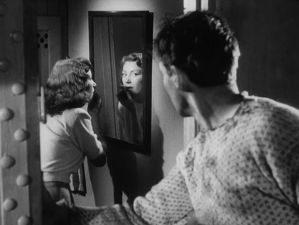 A Ship Bound for India (1947) movie still: medium — A dark, cramped interior space with a wall-mounted mirror reflecting a woman's face. A man in…