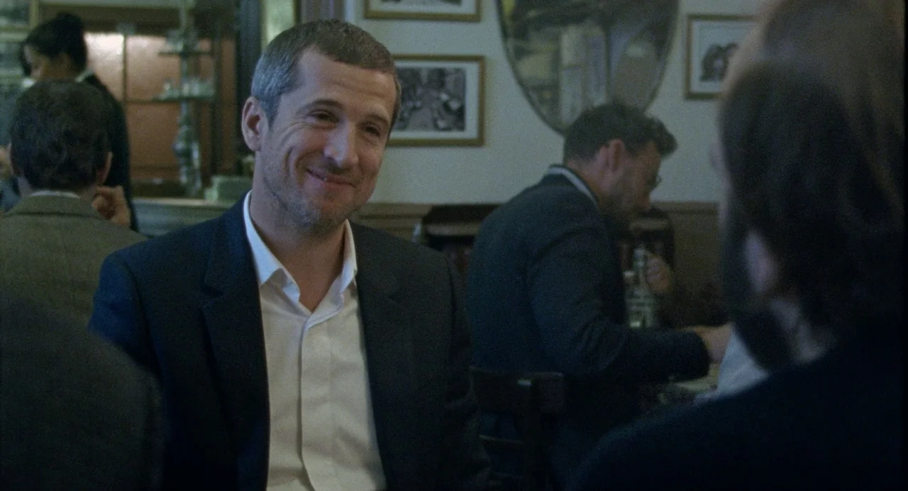 Non-Fiction (2018) movie still: medium — A French bistro interior features framed photographs on the wall and wooden chairs. A man in a dark blazer…