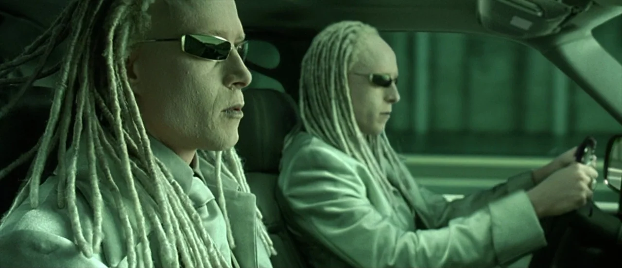 The Matrix Reloaded (2003) movie still: medium — Two identical men with long blonde dreadlocks sit inside the cabin of a car. The pair wear matching…