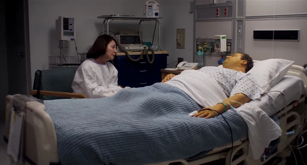 Dark Horse (2011) movie still: medium — A clinical hospital room contains medical monitoring equipment and a bed with a patient. A woman in a gown…