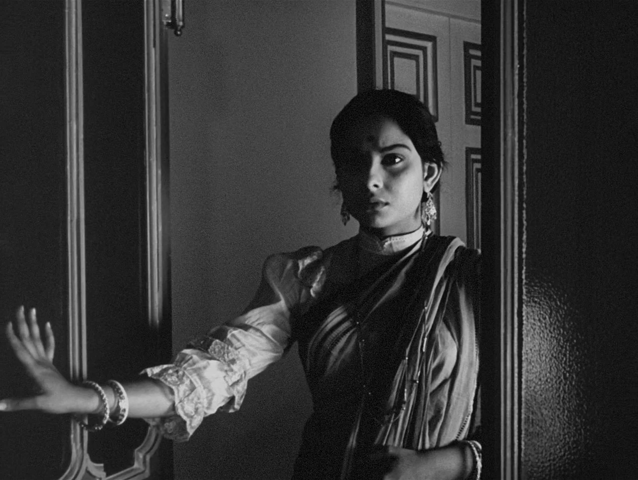Charulata (1964) movie still: medium — A dark indoor space defined by a wooden door frame and architectural molding. A young woman stands in the…