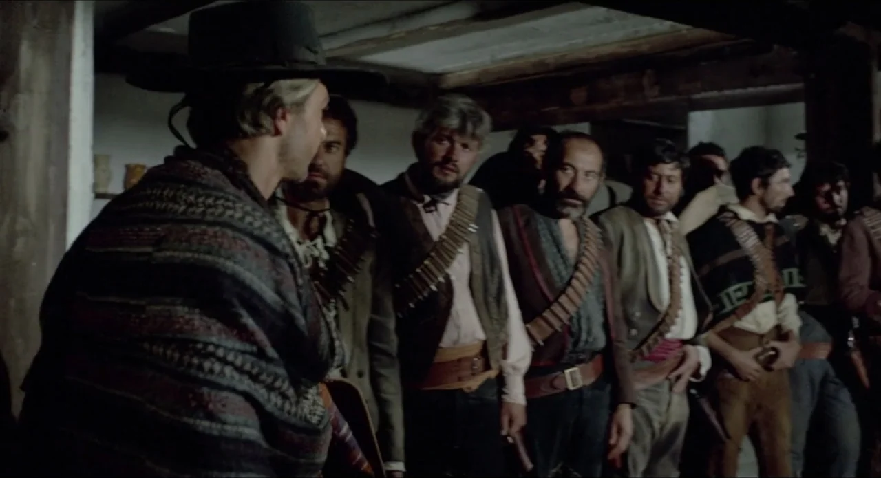 A Stranger in Town (1943) movie still: medium — A dark, wood-beamed interior with a group of men standing in a line. A man in a poncho stands in the…