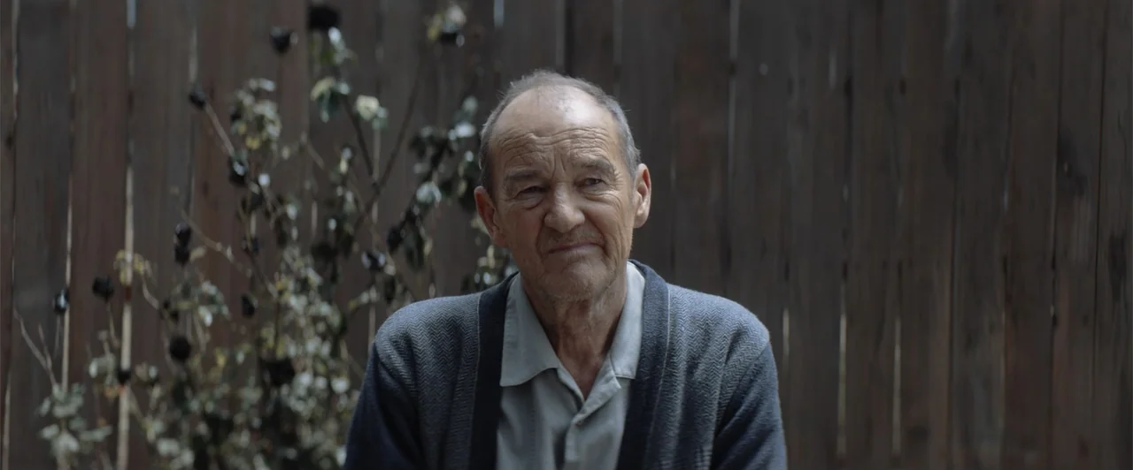 My Neighbor Adolf (2022) movie still: medium — A man sits in front of a dark vertical wood fence with dried foliage. The man wears a cardigan and…