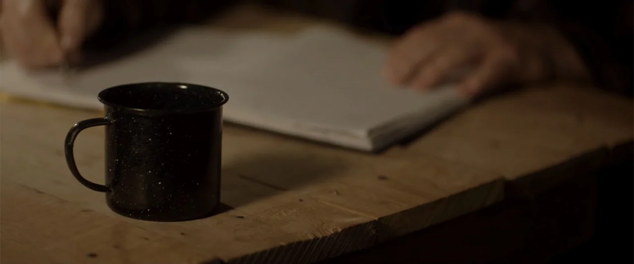 Radius (2017) movie still: closeup — A dark, rustic wooden table surface with a dark speckled mug in the foreground. An out-of-focus person writes…