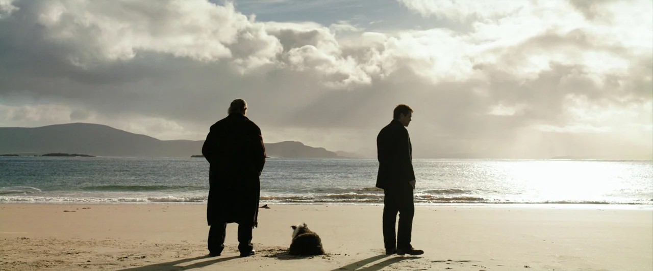 The Banshees of Inisherin (2022) movie still: wide — An overcast beach landscape features distant hills across the water under a cloudy sky. Two men…