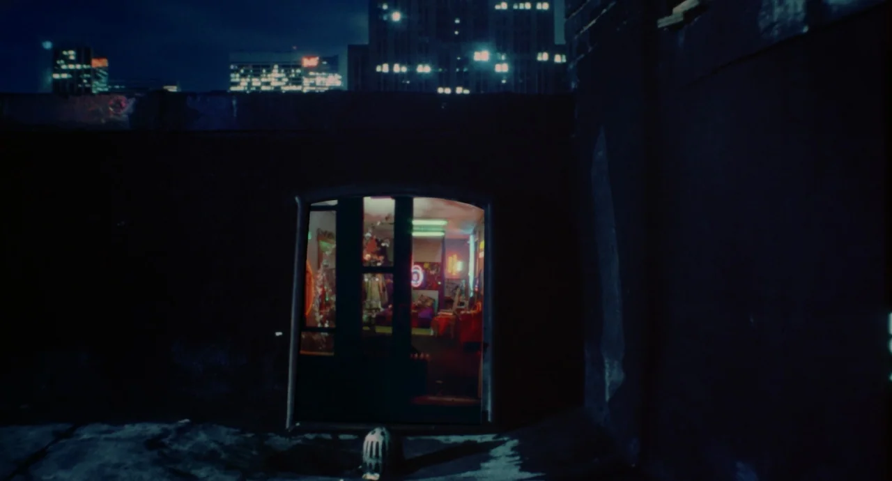 Liquid Sky (1982) movie still: wide — A dark, shadowed urban exterior features a single illuminated doorway leading into a cluttered interior.…
