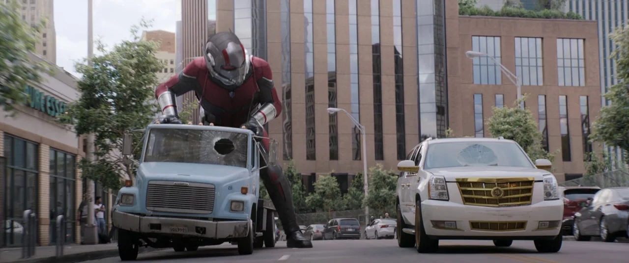 Ant-Man and the Wasp (2018) movie still: wide — A city street features tall office buildings and parked cars along the asphalt road. A giant man in…