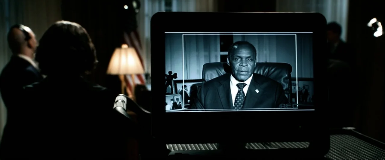 2012 (2009) movie still: medium — A dimly lit room contains a video monitor displaying a man in a formal suit. An observer in the foreground watches…