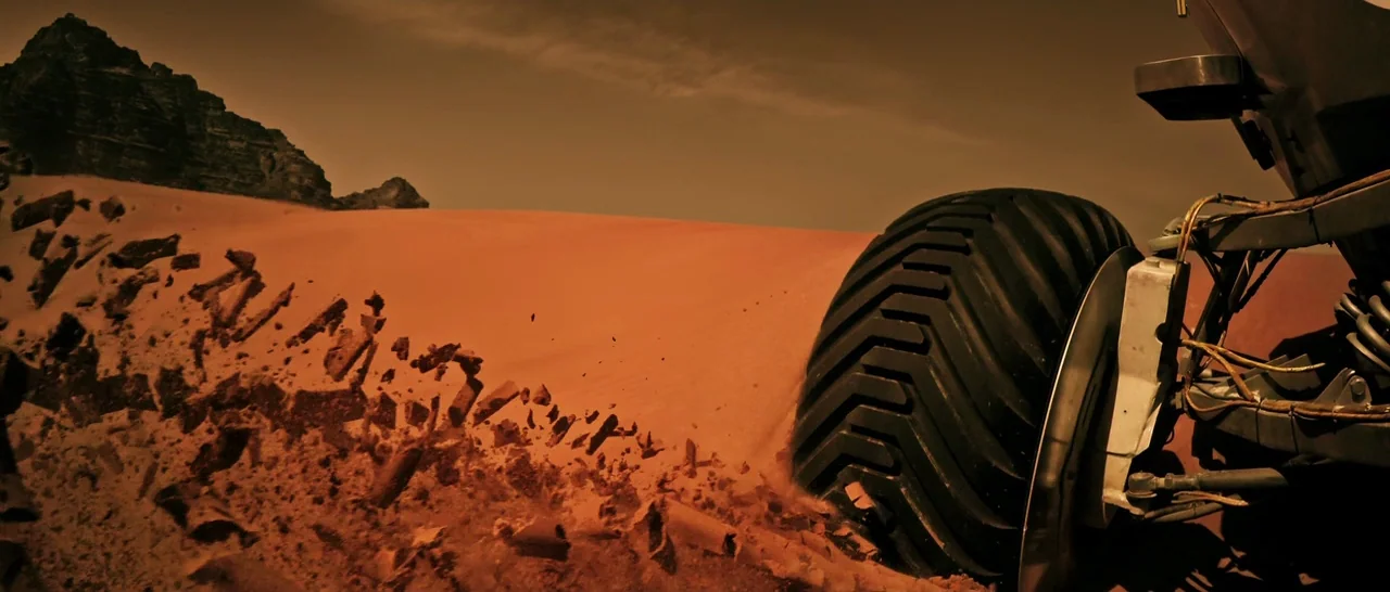 The Martian (2015) movie still: closeup — A vast, arid desert landscape features a rocky mountain peak in the distance. A heavy mechanical vehicle…