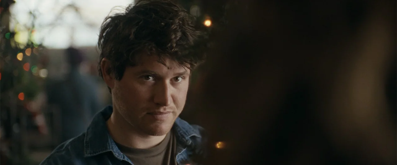 Silent Night, Deadly Night (2025) movie still: closeup — A man stands in a dimly lit indoor space with blurred festive lights nearby. The subject…