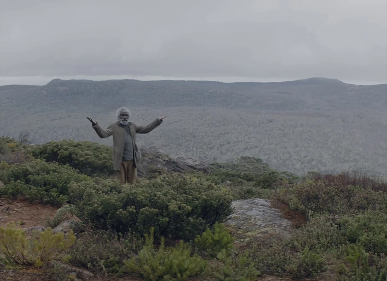 The Nightingale (2018) movie still: fullbody — An elderly man stands on a rocky mountain ridge covered in low green brush. The man has both arms…