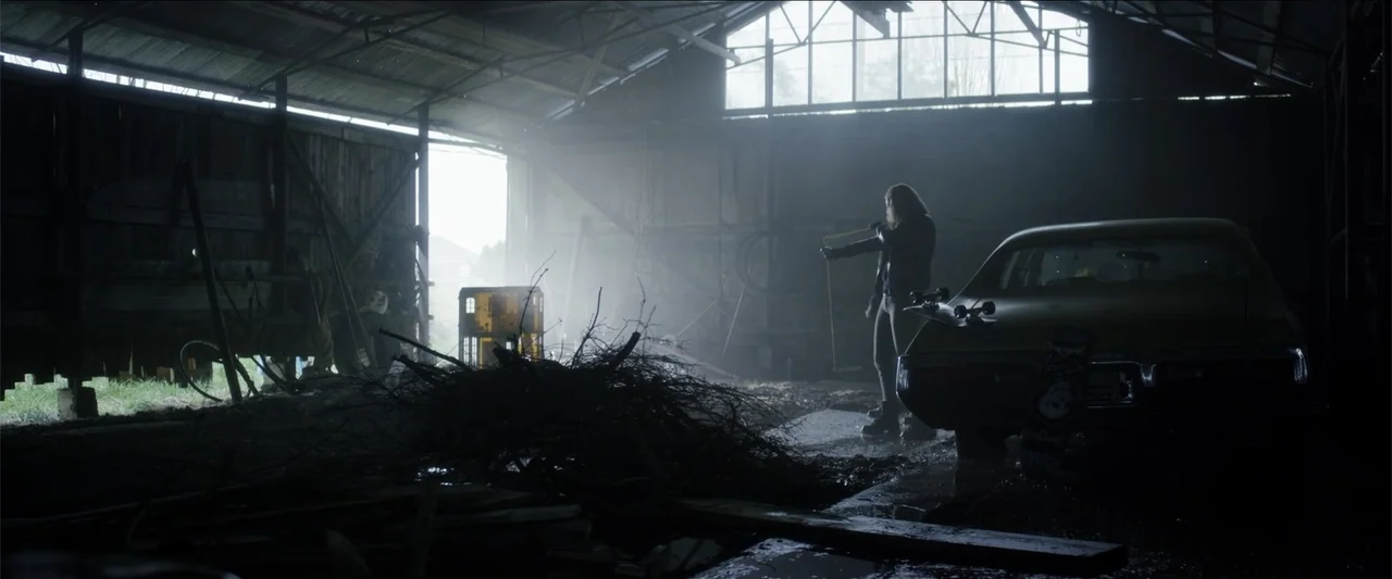 The Hollow Child (2018) movie still: wide — A dark wooden barn contains a classic car and piles of brush debris. A young woman holds a slingshot…