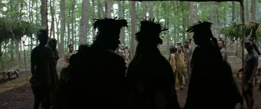 The New World (2005) movie still: fullbody — A group of people stand in a shaded clearing within a dense deciduous woodland. They appear to be in an…