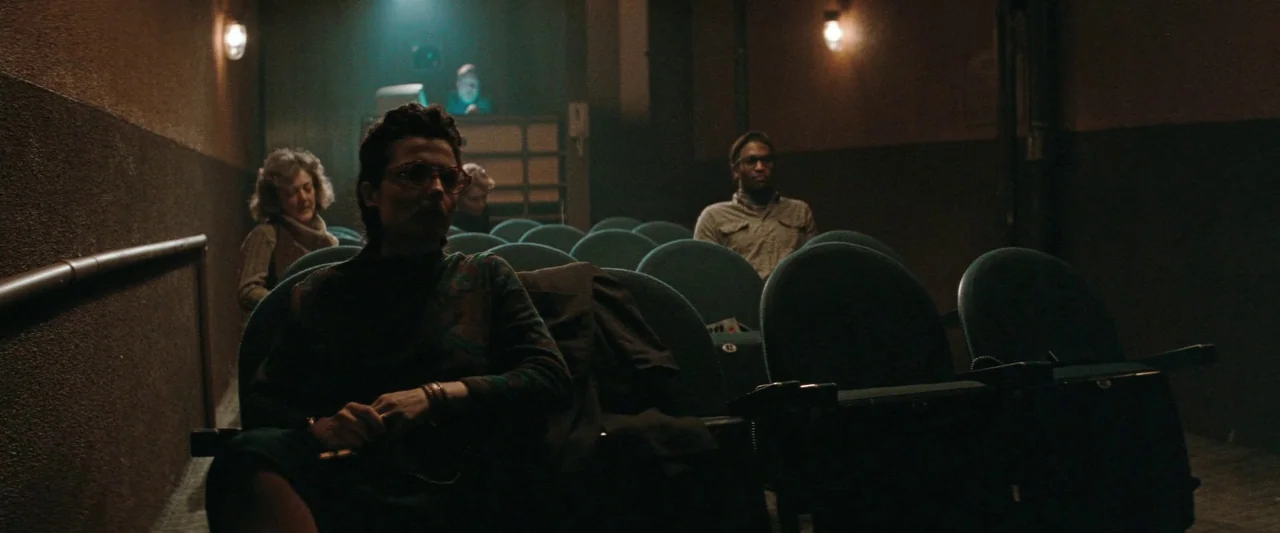 The Adjuster (1991) movie still: medium — A dimly lit movie theater with rows of empty blue velvet seats. Several people sit scattered throughout…