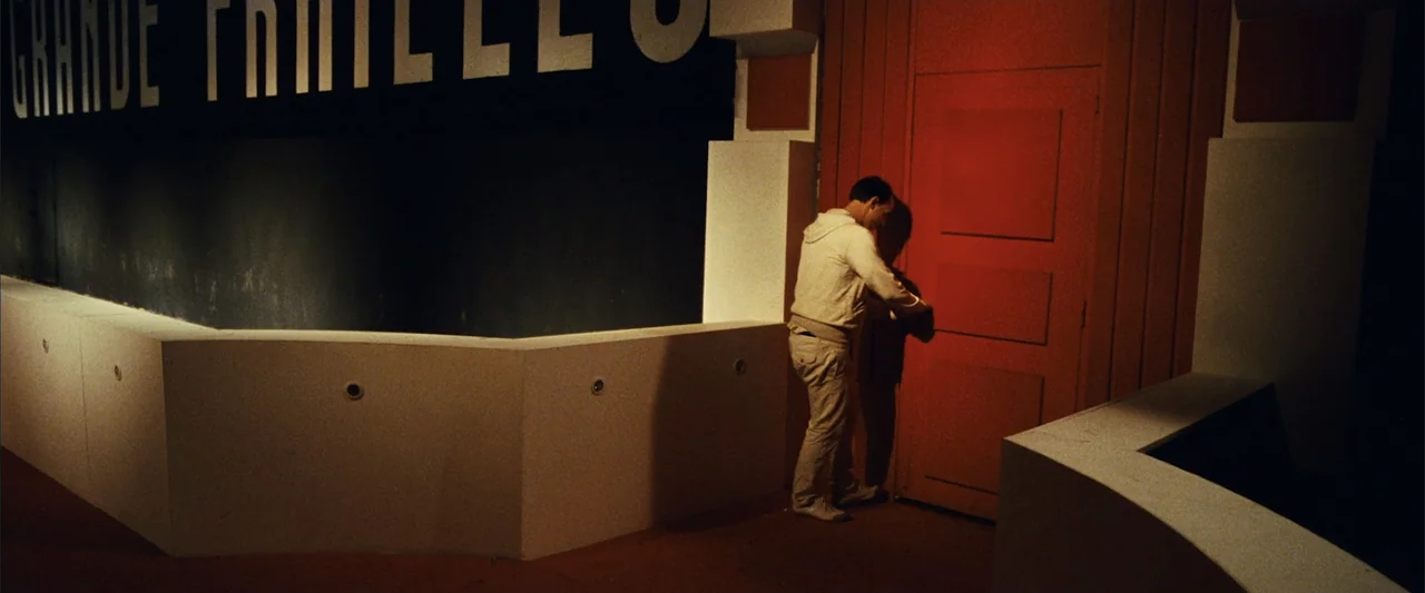 Reality (2023) movie still: fullbody — A modern building interior features bold black wall lettering and a prominent red door. A man in beige…