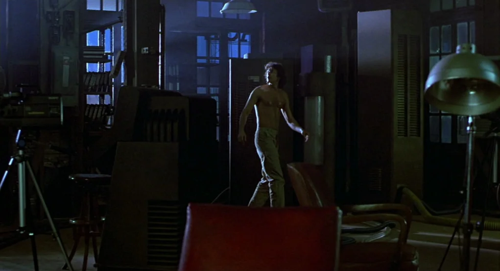 The Fly (1986) movie still: wide — A cluttered production studio features multiple cameras on tripods and dark metal equipment cabinets. A shirtless…