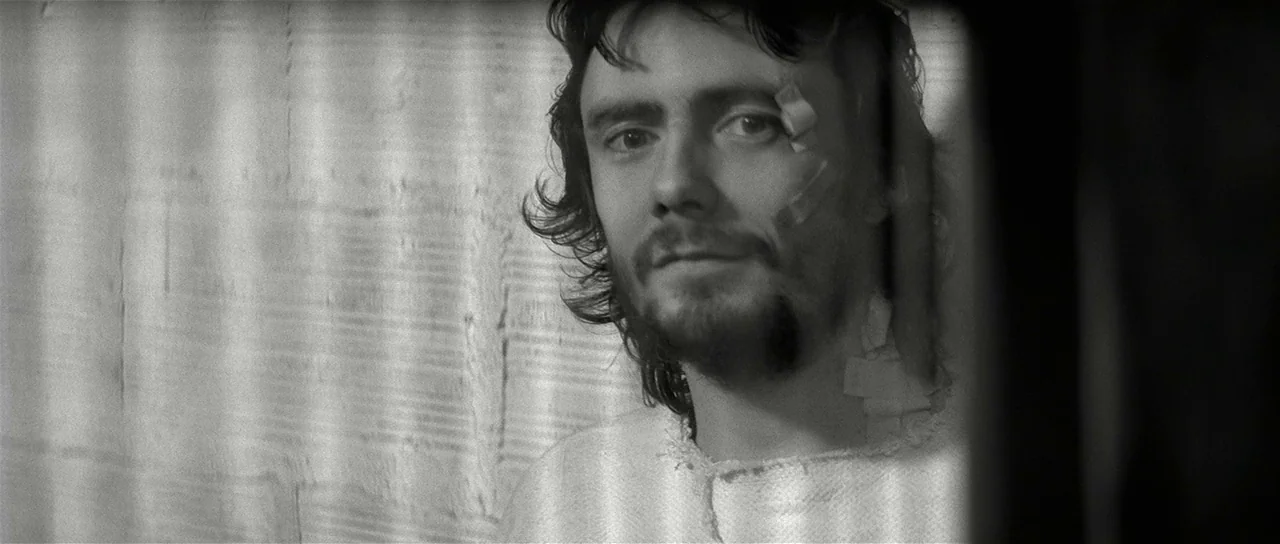 The Last Battle (1983) movie still: closeup — A man stands behind thin vertical bars casting sharp light and shadow on his face. He has a bandage on…