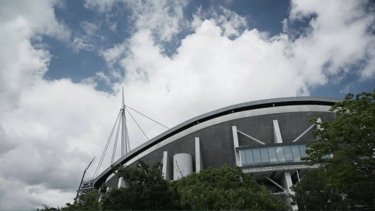 Vai Corinthians (2023) movie still: establishing — A modern stadium exterior features dark stone cladding and a large structural spire. The building…
