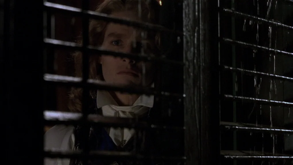 Interview With The Vampire movie still: closeup — A man peers through closed wooden window blinds into the darkness outside at night. Rain streams…