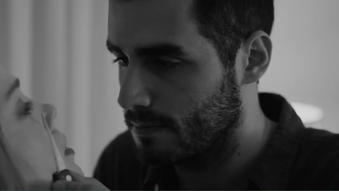 Creditors (2025) movie still: closeup — A dimly lit interior space with a man positioned close to a woman. A man holds a small metal knife near the…