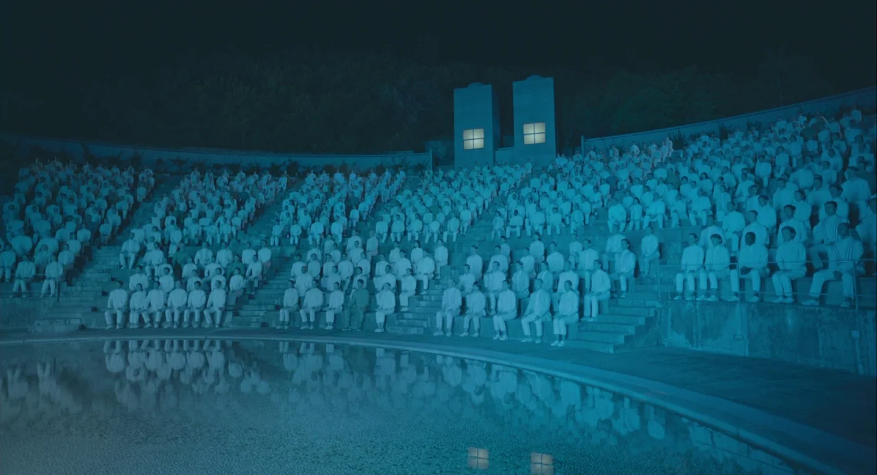 Equals (2015) movie still: wide — A large crowd in white clothing fills the concrete tiers of an amphitheater. A pool of water reflects the seated…