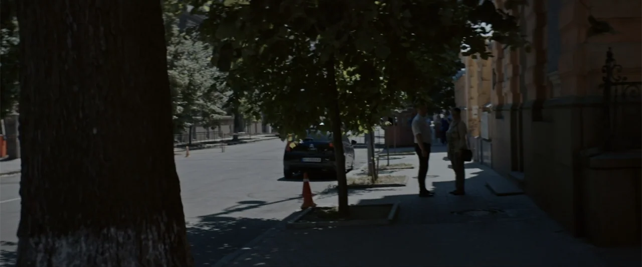 La herencia (2010) movie still: wide — A sunny day on a city sidewalk next to a stone building wall. Two people stand facing each other while a…
