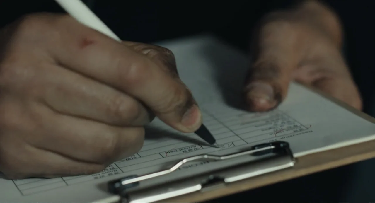 Winter in Sokcho (2025) movie still: closeup — A close-up view of a person filling out a registration form on a clipboard. The hand holding a black…