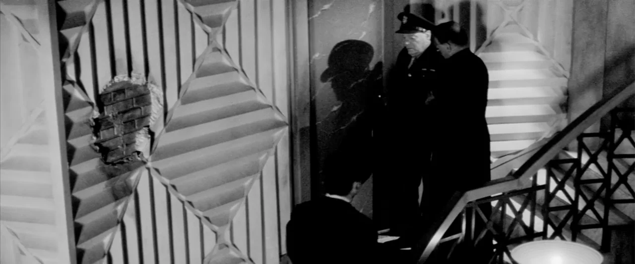 Europa (1991) movie still: medium — An interior staircase features modern geometric wall panels and a jagged hole in masonry. Two men in suits stand…
