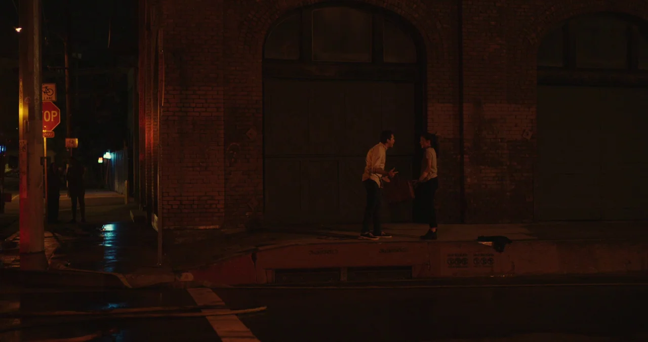 Violet (2021) (2021) movie still: wide — A dark nighttime street corner features a red brick building with arched entryways. A man and a woman stand…