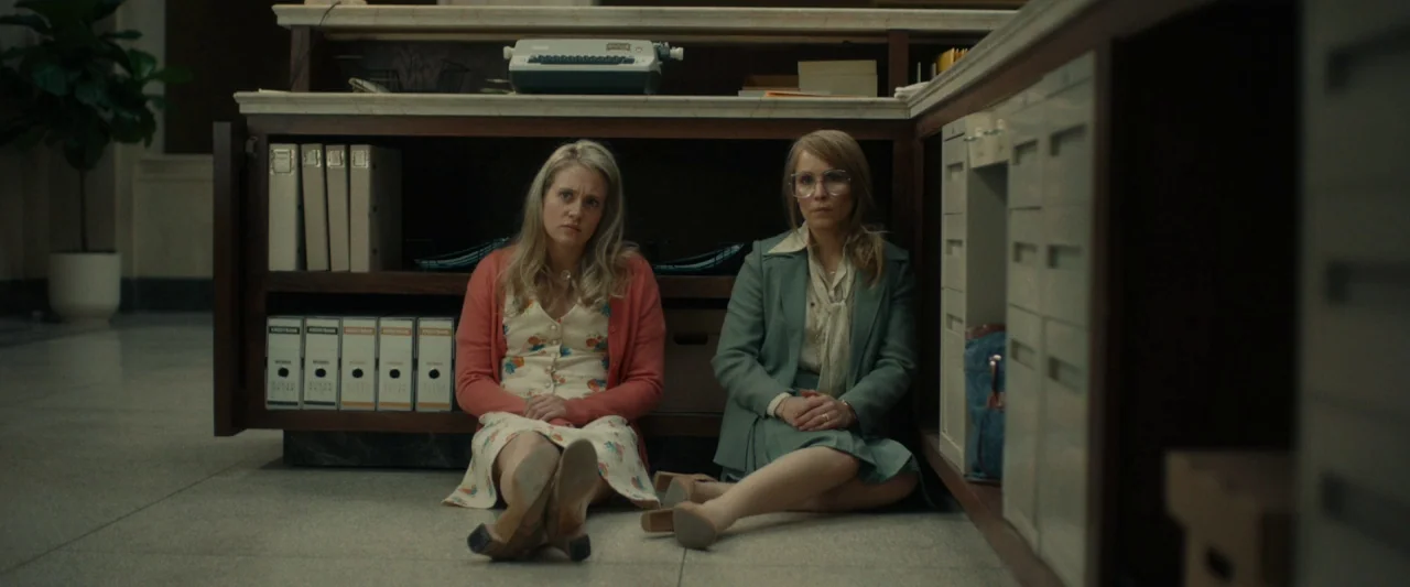 Stockholm (2019) movie still: medium — Two women sit on the linoleum floor behind a wooden counter in a bank. Files, a vintage typewriter, and a…