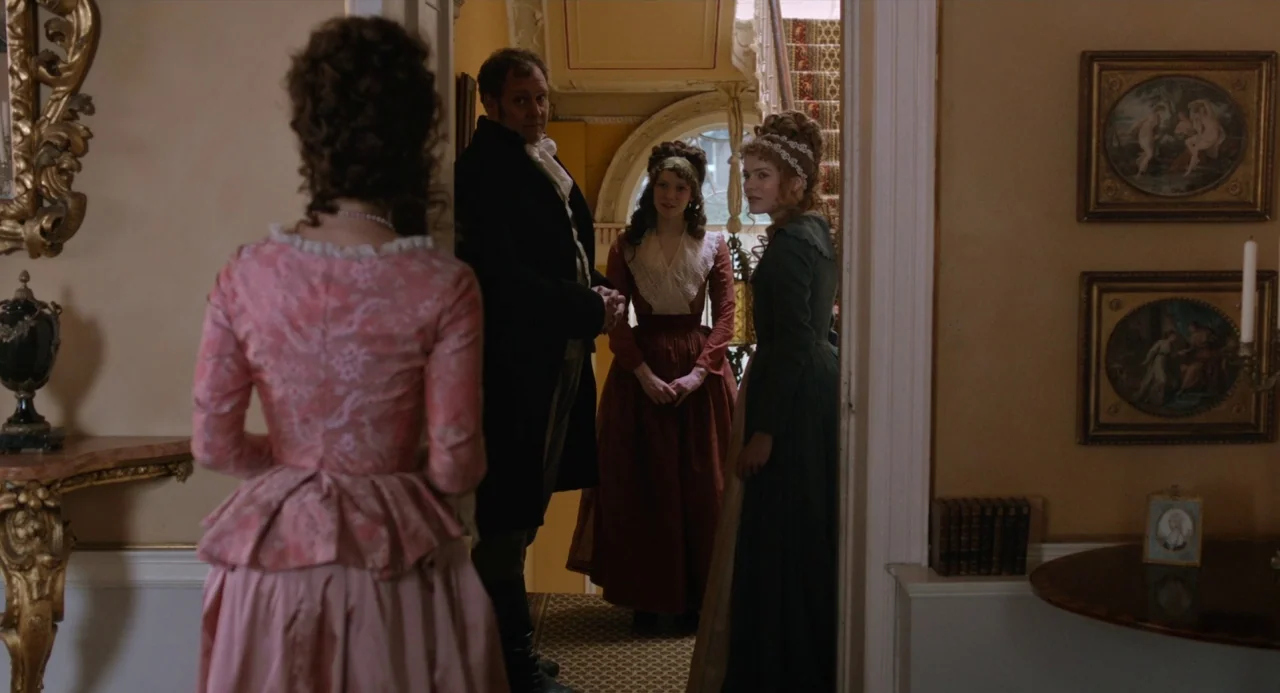 Love and Friendship (2016) movie still: medium — An 1800s English manor entryway features warm yellow walls and ornate gold decor. A woman in pink…