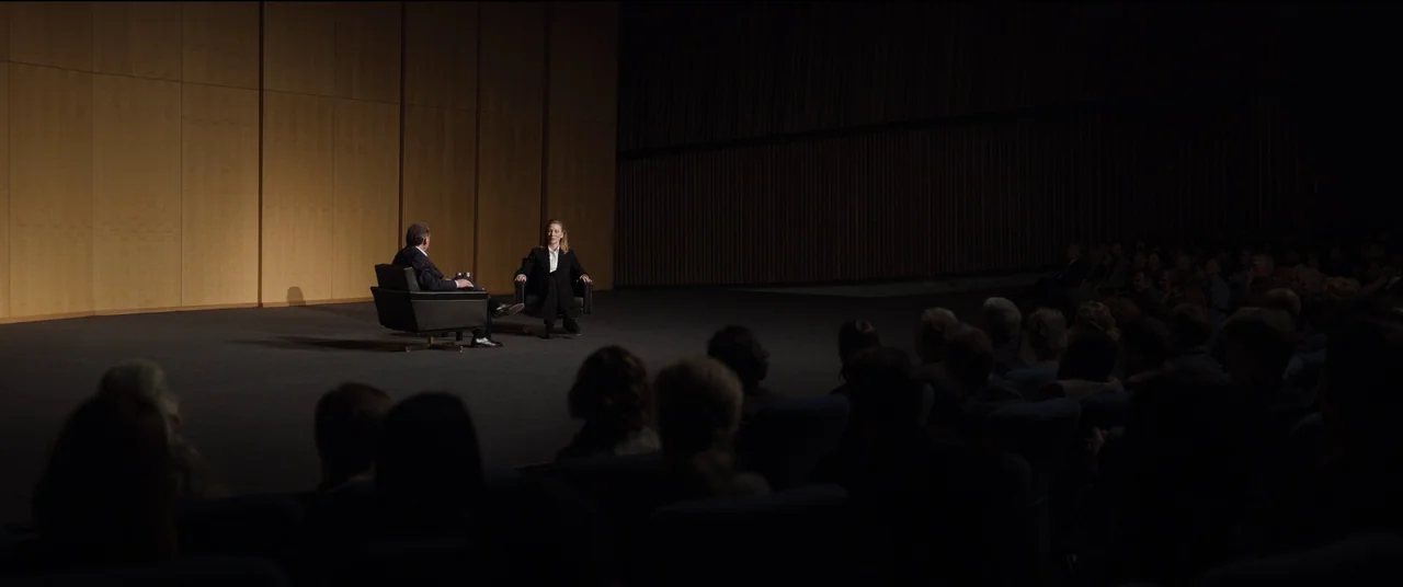 Tár (2022) movie still: wide — A spacious auditorium features a stage with wood-paneled walls and two black leather chairs. A man and a woman sit on…