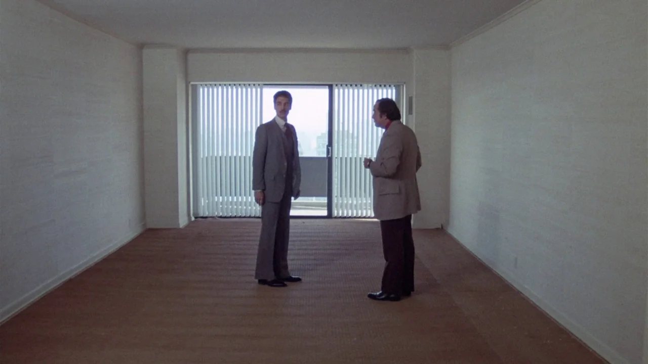 The Sentinel (1977) movie still: fullbody — An empty apartment space features white walls, tan carpeting, and a large sliding door. Two men in suits…