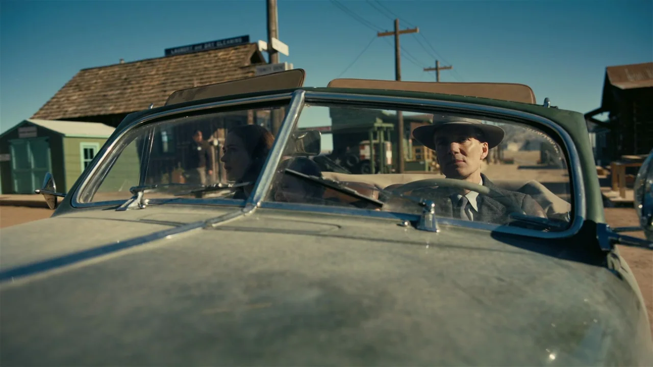 Oppenheimer (2023) movie still: medium — A man and woman sit inside a dusty vintage convertible parked in a desert town. Wooden buildings and…