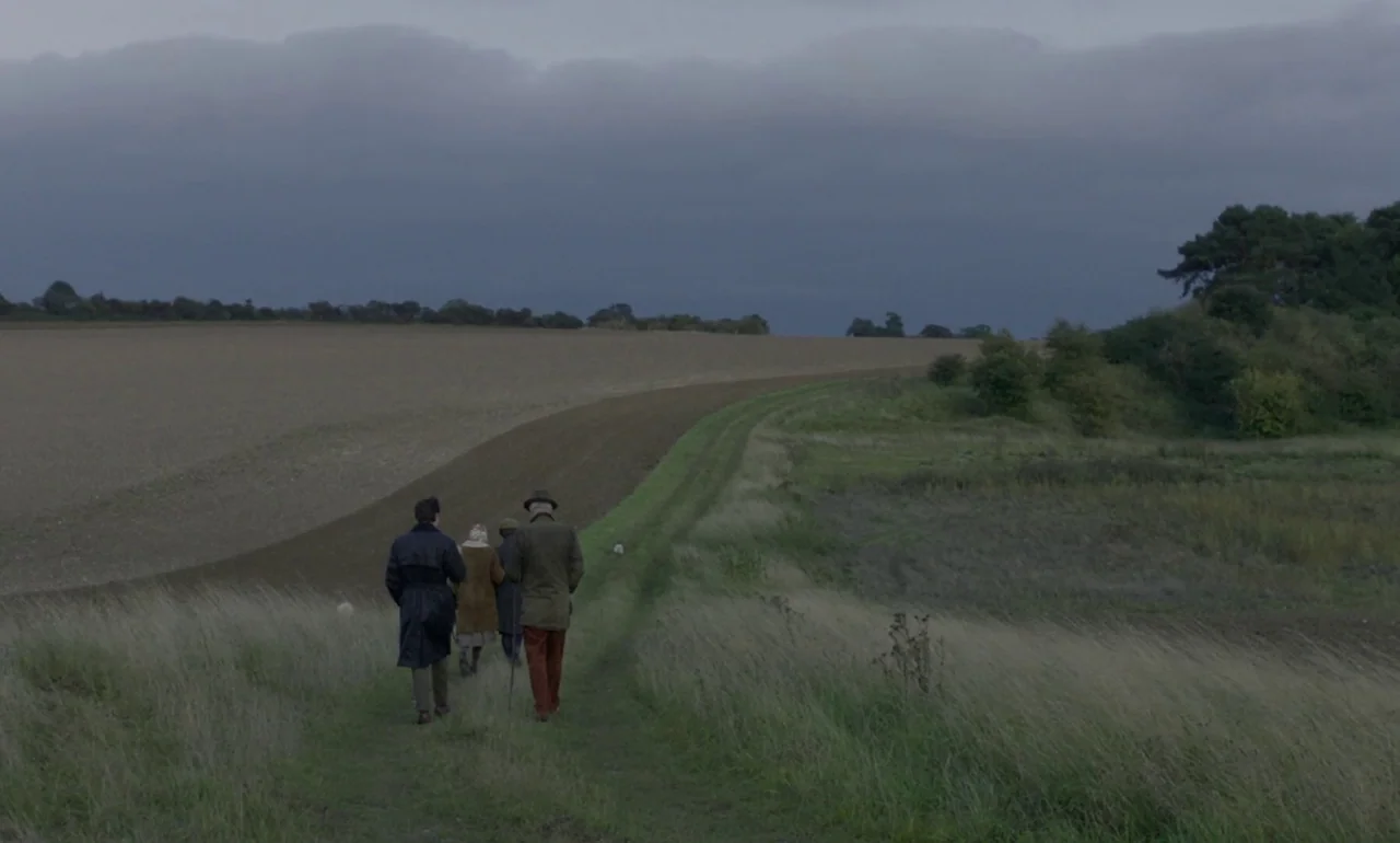 The Souvenir (2019) movie still: wide — A group of people walk along a dirt path bordering a large ploughed field. Low heavy grey clouds hang over…