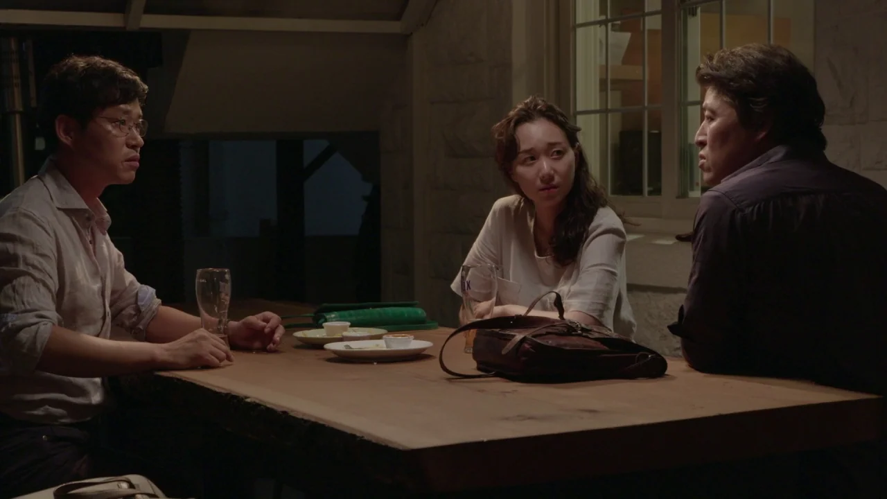Yourself and Yours (2016) movie still: medium — A dimly lit outdoor seating area features a long wooden table at night. Three people sit around the…
