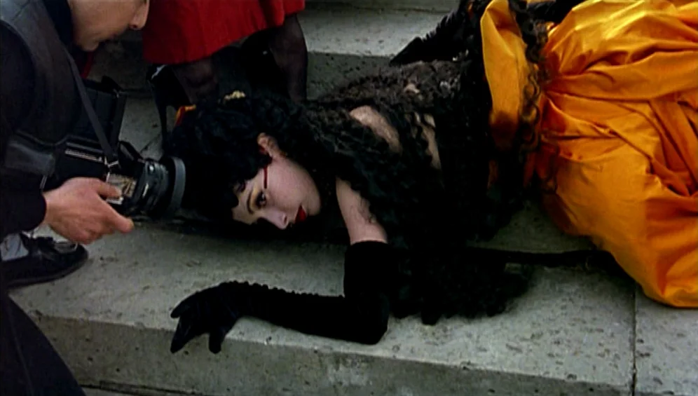 The Tango Lesson (1997) movie still: medium — A woman in an orange skirt lies on concrete steps outdoors. A photographer holds a camera close to her…