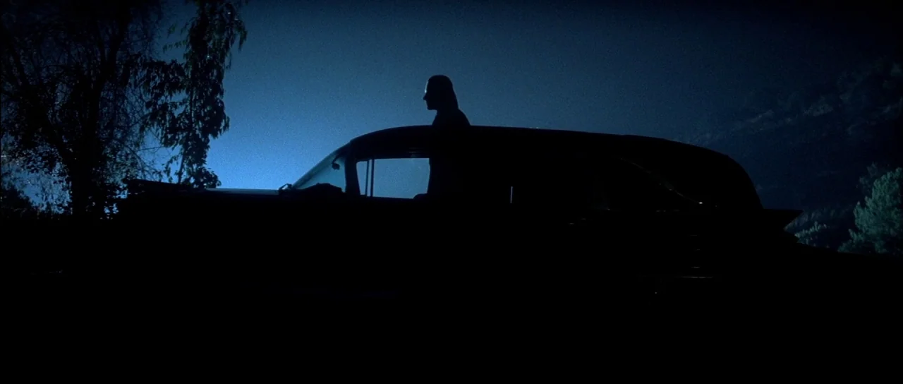 The Monster Squad (1987) movie still: wide — A dark night landscape featuring a silhouette of a car against a blue sky. A man stands upright through…