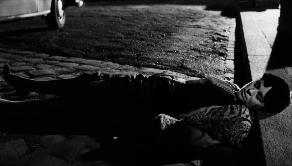 Veronika Voss (1982) movie still: medium — A woman lies motionless on a dark, wet cobblestone street at night. A car tire is partially visible in…