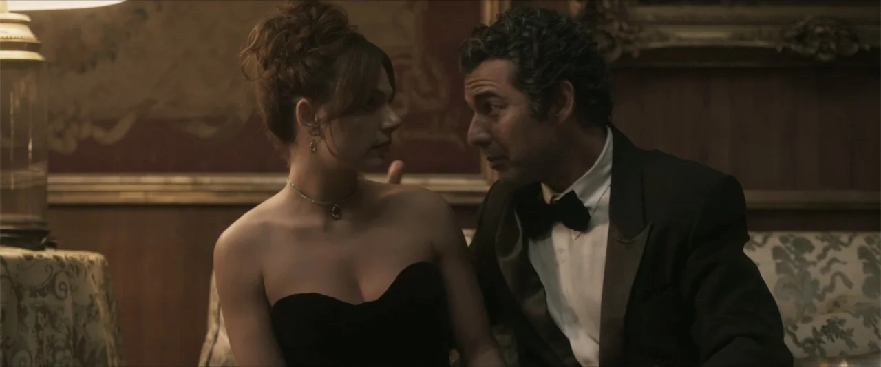 Angela (2023) movie still: medium — An interior space features wood-paneled walls and ornate framed wall hangings. A woman and a man in formal…