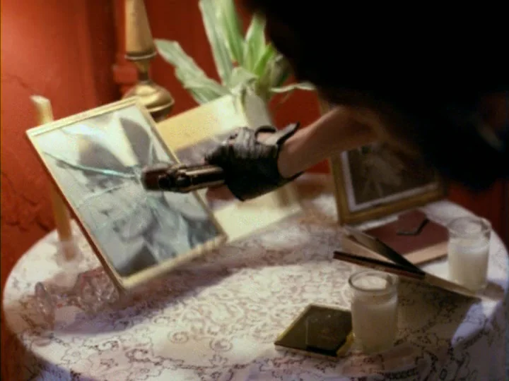 Tenement (1985) movie still: closeup — A small round table draped in a white lace cloth holds framed photographs and candles. A hand wearing a black…
