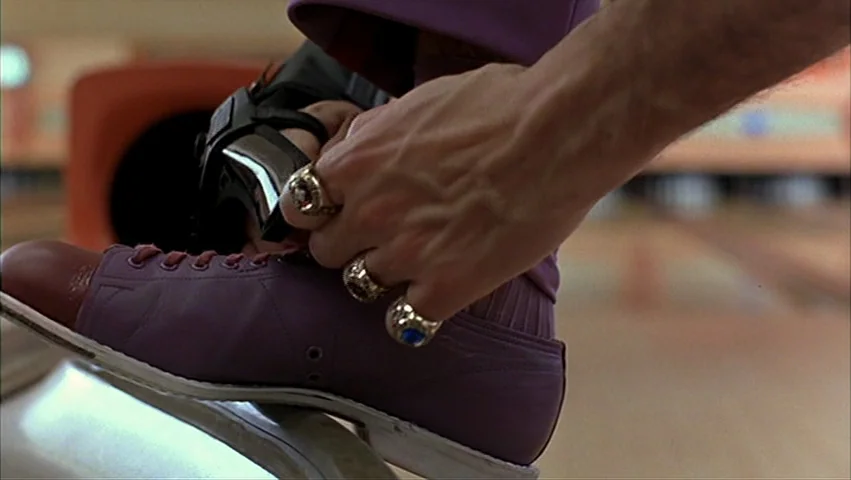The Big Lebowski (1998) movie still: closeup — A close-up of a person tying a bowling glove on their foot. The person is wearing purple bowling…