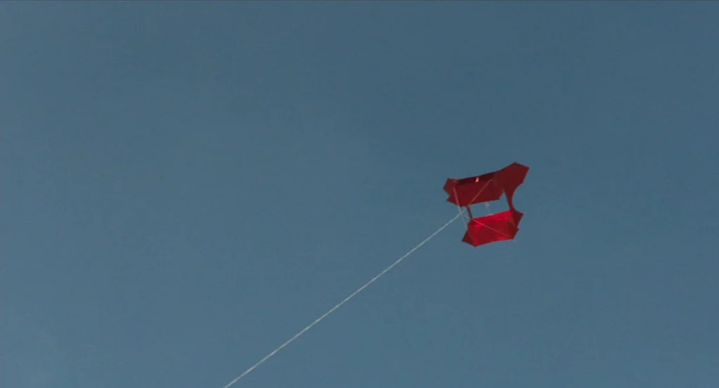 The Congress (2013) movie still: wide — A bright red box kite flies against a clear blue sky. A thin white string connects the kite to an unseen…