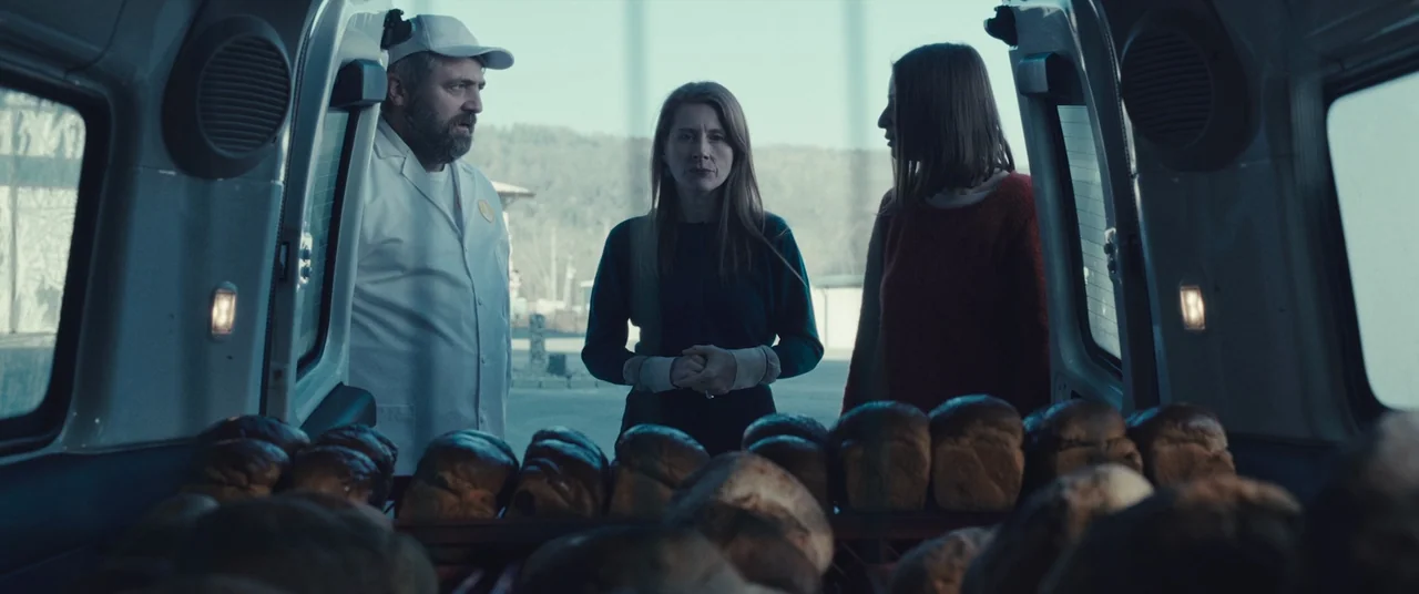 R.M.N. (2022) movie still: medium — The open cargo doors of a van frame three people standing outdoors. Racks of bread loaves fill the immediate…